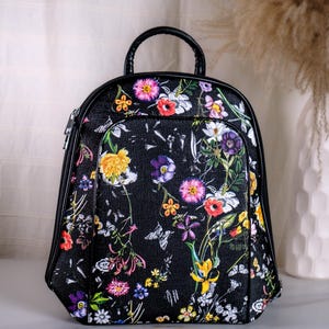 Könnte beinhalten: Ein schwarzer Rucksack mit einem lebendigen, mehrfarbigen Blumenmuster aus verschiedenen Blüten und Schmetterlingen. Die Tasche hat eine abgerundete Oberseite, einen Tragegriff oben und schwarze Kanten, mit einem Hauptfach mit Reißverschluss.