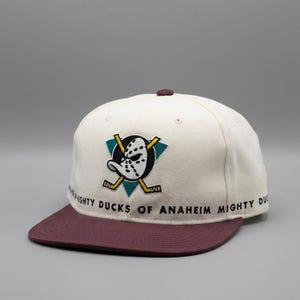Puede incluir: Una gorra de béisbol color blanco hueso con visera y botón superior color granate. El frente presenta un logotipo bordado de los Mighty Ducks of Anaheim con una máscara de portero y palos de hockey cruzados. El texto MIGHTY DUCKS OF ANAHEIM está impreso en la base.