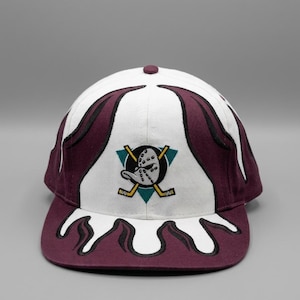 May include: A white and maroon baseball cap featuring a central embroidered logo of a hockey goalie mask shaped like a duck head with two crossed hockey sticks. The cap displays flame-like patterns in maroon and white across the crown and brim.