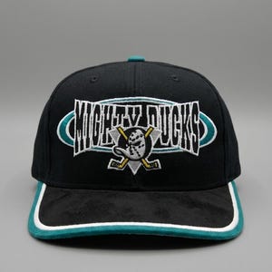 Può includere: Un cappellino da baseball nero con il testo ricamato MIGHTY DUCKS in bianco con un bordo color ottanio. Sotto il testo si trova un logo circolare raffigurante una maschera da portiere e due bastoni da hockey incrociati in bianco, nero e giallo.