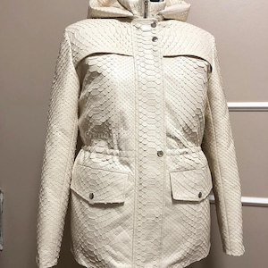 May include: An off-white hooded jacket featuring a textured snakeskin-like pattern throughout. The garment includes a front zip closure concealed by a snap-button placket, two front flap pockets with snap buttons, and a drawstring waist detail.