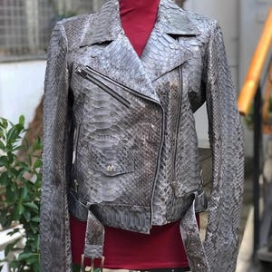 May include: A grey snakeskin-patterned leather motorcycle jacket featuring a notched lapel, an asymmetrical front zip closure, long sleeves, a small flap pocket with a silver-toned snap button, and a matching belt with a metal buckle.
