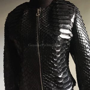 May include: A black jacket featuring a textured, overlapping scale pattern across the entire surface. The garment has a central silver-toned metal zip closure and a round neckline. The text "GenuinePythonJacket" is visible on the front.