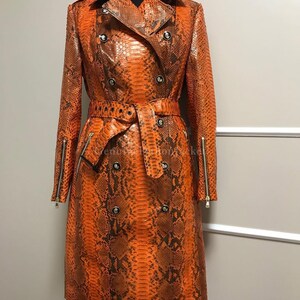 Könnte beinhalten: Ein zweireihiger Trenchcoat mit einem leuchtend orangefarbenen und dunkelbraunen Schlangenledermuster. Der Mantel verfügt über einen passenden Taillengürtel mit Schnalle, ein Revers, silberfarbene Knöpfe und vertikale Reißverschlüsse an den Ärmeln.
