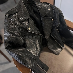 Könnte beinhalten: Eine schwarze Jacke mit texturiertem Schlangenledermuster und metallischem Glanz. Das Kleidungsstück verfügt über ein steigendes Revers, silberfarbene Metalldruckknöpfe und einen Frontreißverschluss. Der Text GenuinePythonJacket ist sichtbar.