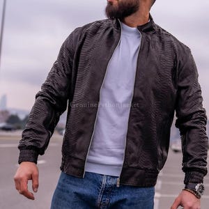 May include: A dark brown bomber jacket with a textured snakeskin pattern, featuring a stand-up collar, a central silver zip, and ribbed cuffs. The jacket is worn over a plain white t-shirt and blue denim jeans. The text GenuinePythonJacket is visible.