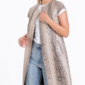 May include: A sleeveless long waistcoat featuring a grey and beige snakeskin pattern, a round neckline, and a vertical row of gold-toned buttons down the front. The garment is worn over a plain white t-shirt and light blue denim jeans with visible seams.