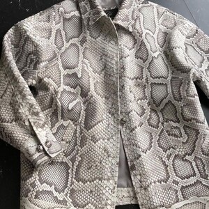 May include: A long-sleeved button-down jacket featuring a grey and white snakeskin pattern. The garment has a standard collar, a front button closure with metallic buttons, and buttoned cuffs. The interior is lined with a solid grey fabric.