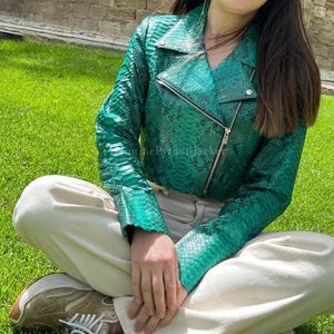 May include: A vibrant emerald green motorcycle jacket featuring a textured snakeskin pattern, a diagonal silver-tone metal zip closure, notched lapels with silver-tone snap buttons, and long sleeves. The jacket is worn with cream-coloured trousers.