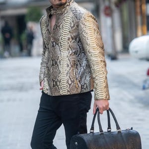 Op de afbeelding: Een jas met lange mouwen en een gedetailleerd slangenleerpatroon in beige, bruine en crèmetinten. De jas heeft een hoge kraag met een drukknoopsluiting, een ritssluiting aan de voorzijde en verticale zakken met rits aan de zijkanten.