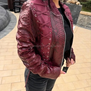 May include: A burgundy jacket featuring a textured snakeskin pattern on the sleeves and back, with a smooth solid-coloured front panel accented by silver-toned metal eyelets and crisscross lacing. The jacket includes a central zip and a ribbed collar.