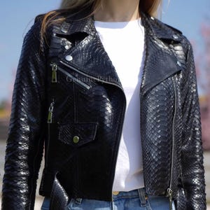 May include: A black motorcycle jacket featuring a textured snakeskin pattern, a wide notched collar, silver-toned metal zips, and a small flap pocket on the front. The jacket is worn over a plain white t-shirt and blue denim jeans.