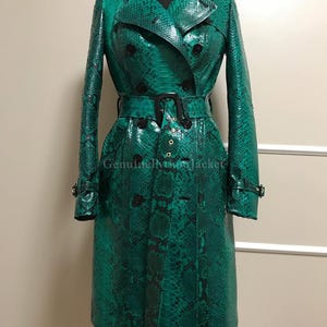May include: A vibrant emerald green trench coat featuring a distinct snakeskin pattern, a double-breasted front with black buttons, a matching waist belt with a black buckle, and epaulets on the shoulders. The text GenuinePythonJacket is visible.