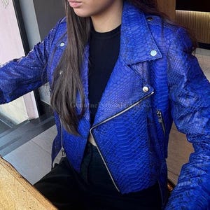Könnte beinhalten: Eine leuchtend blaue Lederjacke mit Schlangenmuster. Die Jacke hat einen klassischen Biker-Stil mit Reverskragen, asymmetrischem Reißverschluss und silberfarbenen Beschlägen. Ein auffälliges Kleidungsstück.