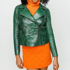 May include: A green snakeskin-effect leather jacket with a black zip. The jacket has a classic biker style with a notched collar and long sleeves. The model is wearing an orange turtleneck and orange mini skirt.
