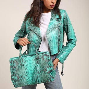 May include: A turquoise and black snakeskin-patterned leather handbag with a matching tassel, held by a person wearing a turquoise snakeskin-patterned leather jacket. The jacket has a black zipper and a classic biker style.