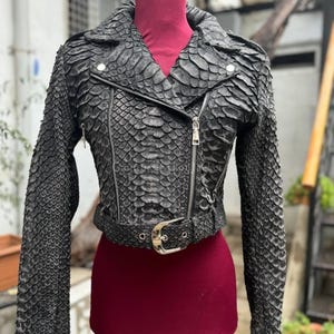 Puede incluir: Una chaqueta estilo motero de color negro con un patrón texturizado similar a escamas en toda la superficie. La chaqueta incluye un cuello con solapas, un cierre de cremallera asimétrico de metal plateado, mangas largas y un cinturón a juego con hebilla plateada.