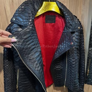 Könnte beinhalten: Eine schwarze Jacke mit einer strukturierten Schuppenoptik auf der Außenseite, einem leuchtend roten Innenfutter und goldfarbenen Metallreißverschlüssen. Das Kleidungsstück hat einen breiten Kragen mit silbernen Druckknöpfen und hängt an einem goldenen Kleiderbügel.