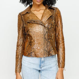 May include: A brown and tan snakeskin-patterned leather jacket featuring a notched collar, long sleeves, and multiple gold-tone metal zips. The jacket has an asymmetrical front zip closure, two zipped chest pockets, and two zipped side pockets.