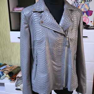 May include: A grey jacket with a textured snakeskin pattern, featuring a notched lapel, an asymmetrical silver-tone zip closure, and two zipped side pockets. The garment is displayed on a black mannequin torso.