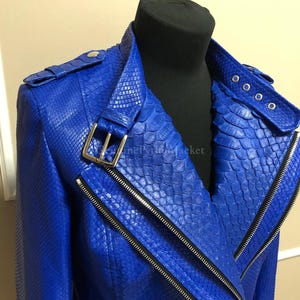 Könnte beinhalten: Eine leuchtend blaue Jacke mit strukturiertem Schlangenhautmuster, einem breiten Kragen mit silbernem Schnallenriemen und einem asymmetrischen Frontreißverschluss. Das Kleidungsstück verfügt über Schulterklappen mit silbernen Beschlägen.
