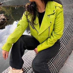 May include: A bright lime green biker jacket featuring a textured snakeskin pattern, silver-toned metal zips, and snap button lapels. The jacket is worn over a black top and paired with dark charcoal grey denim jeans, creating a bold look.