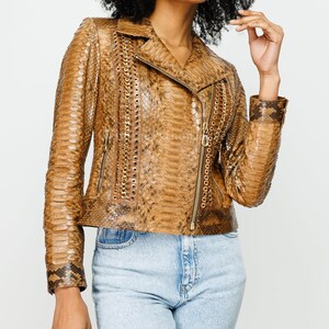 Könnte beinhalten: Eine Jacke mit Schlangenledermuster in Beige und Braun, ausgestattet mit einem Reverskragen, einem asymmetrischen goldenen Reißverschluss sowie vertikalen Einsätzen, die mit goldenen Metallösen und eingearbeiteten Ketten verziert sind.