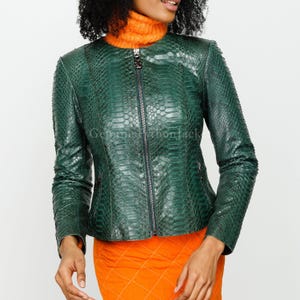 May include: A dark green jacket with a snakeskin texture, featuring a round neckline, long sleeves, and a central silver zip closure. The jacket is worn over an orange turtleneck jumper and an orange quilted skirt, with a visible watermark.