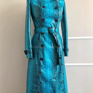 Op de afbeelding: Een lange, dubbelrijige trenchcoat met een levendig turquoise en zwart slangenleerpatroon. De jas heeft een kraag, lange mouwen met gespmanchetten, een bijpassende ceintuur met gesp en donkere knopen. De tekst luidt Genuine Python Japan.