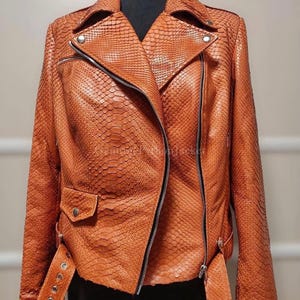 May include: A burnt orange motorcycle jacket featuring a textured snakeskin pattern, a wide notched collar, silver-toned metal zips, and a buckled belt at the hem. The jacket includes a diagonal front zip closure and a small flap pocket.
