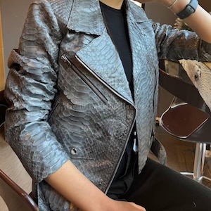 May include: A gray snakeskin-textured leather jacket featuring a notched lapel, long sleeves, and a diagonal silver-tone metal zipper closure. The jacket includes a small zippered pocket on the chest and a flap pocket with a silver button detail.