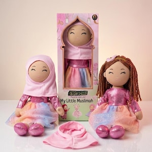 Könnte beinhalten: Zwei Plüschpuppen mit brauner Haut, die rosa Hijabs und glänzende rosa Langarm-Oberteile mit mehrfarbigen Glitzer-Röcken tragen. Eine Puppe hat braune Garnhaare, die andere trägt einen rosa Hijab. Eine Schachtel mit der Aufschrift ist dabei.