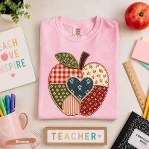Puede incluir: Una camiseta rosa claro con una gran aplicación en forma de manzana hecha de varios parches de tela en rojo, verde y crema con patrones florales, de cuadros y de rayas. Un parche en forma de corazón azul oscuro con detalles florales blancos.