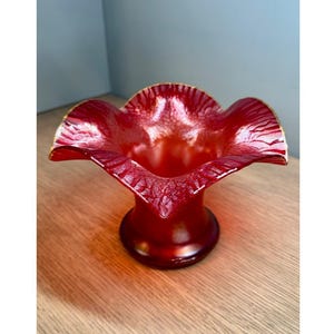 May include: A vibrant red glass vase featuring a wide, ruffled, and crimped rim with a textured, crackled finish. The body of the vase tapers down to a smooth, circular base, showcasing a deep crimson hue that transitions to a lighter, translucent red.