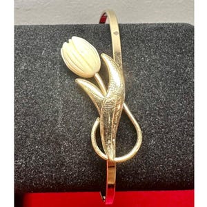 May include: Gold-tone bracelet featuring a tulip design. The bracelet has a slender band and a detailed tulip with a cream-colored flower and gold-colored stem and leaves. The bracelet is a floral-themed accessory.
