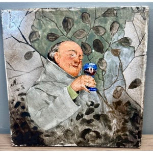 May include: A square ceramic tile featuring a painted illustration of a bald individual wearing a light gray hooded robe. The figure holds a small blue goblet filled with red liquid. The background depicts stylized dark green foliage and branches.