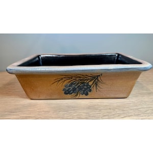 May include: A rectangular ceramic dish with a light brown exterior and a black interior. The dish features a hand-painted design of a pine branch with blue pinecones. The rim is accented with a blue-gray trim. The dish is sitting on a wooden surface.