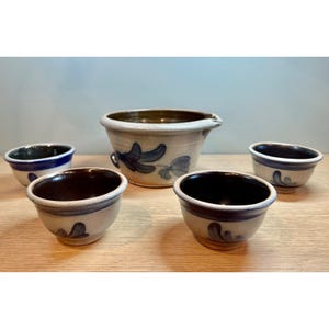 May include: A set of handmade pottery bowls and a pitcher. The bowls have a dark brown interior and a light beige exterior with blue floral designs. The pitcher has a spout and a similar design. The pottery is arranged on a wooden surface.