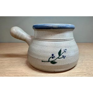 May include: A beige ceramic soup bowl with a handle and a blue rim. The bowl features a green and blue floral design. The bowl is sitting on a wooden surface.