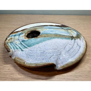 May include: A flat, circular ceramic flower frog with a glazed finish in shades of blue, white, and brown. The surface features a speckled texture and a central circular opening designed to hold flower stems, with a dark brown rim edge.
