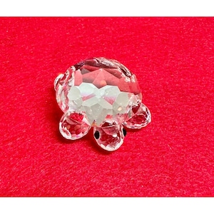 May include: A clear crystal turtle figurine with faceted surfaces. The turtle has a rounded body, four flipper-like legs, and a small head with black eyes. The figurine is set against a red background.