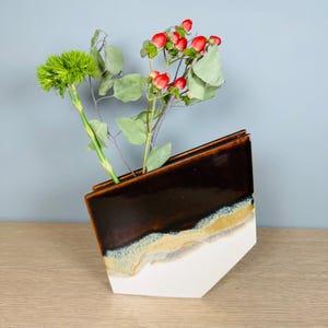 May include: A geometric ceramic vase with a slanted top edge, featuring a glossy dark brown upper section that transitions into a textured beige and blue-grey band, ending in a matte white base, holding green stems and red berries.
