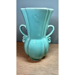 May include: A turquoise ceramic vase with a glossy finish, featuring a wide, flared rim, a bulbous body with vertical fluted panels, and two symmetrical curved handles on the sides. The vase has a smooth, solid color surface throughout.