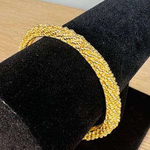 May include: A gold-toned bangle bracelet featuring a textured, beaded surface with a repeating pattern of small, rounded metallic spheres. The circular jewelry piece has a consistent width and a reflective finish across its entire exterior surface.