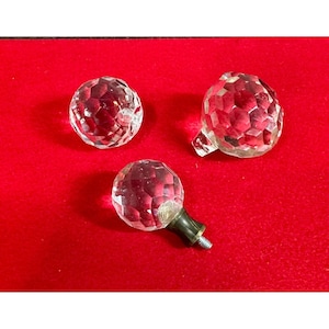 May include: Three clear, faceted crystal knobs on a red surface. One knob has a brass-colored base with a screw. The other two knobs are spherical with a cut-glass design. These decorative knobs could be used for furniture or other craft projects.