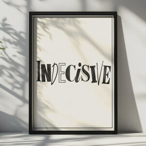 May include: A rectangular wall art print in a black frame featuring the word INDECISIVE in a stylised, mixed-font black typeface on a cream-coloured background. The letters vary in thickness and style, creating a distinct visual contrast.