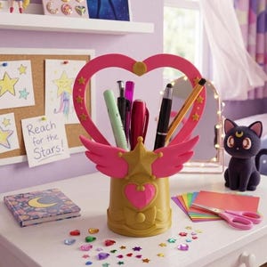 May include: A gold and pink pen holder with a heart-shaped top and wings, holding pens and pencils. The desk also features a black cat figurine, a notebook, and colorful craft supplies. A sign says "Reach for the Stars!"