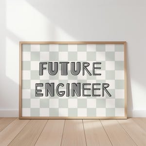 Op de afbeelding: Een ingelijste print met de tekst "FUTURE ENGINEER" in een vetgedrukte, omlijnde lettertype. De achtergrond is een dambordpatroon van lichtgrijze en witte vierkanten. De lijst is licht van hout, en de print staat op een houten vloer.
