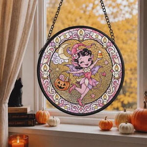 Può includere: Un acchiappasole circolare in vetro colorato raffigurante una fata rosa con ali, che tiene in mano una caramella a forma di mais e un secchiello a forma di zucca. Il design include una mezzaluna, stelle e candele accese entro un bordo decorativo, appeso a una catena.