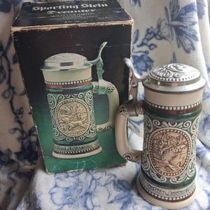 May include: A ceramic beer stein with a metal lid and handle, featuring a relief of a rainbow trout and the text The Strike, Rainbow Trout. Beside it is a black box labeled Sporting Stein Decanter, Wild Country Cologne, 8 FL. OZ.
